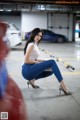 A woman in a white top and blue jeans crouching down in a parking garage.