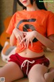 A woman in an orange shirt and red shorts sitting on the floor.