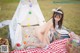 A naked woman sitting on a picnic blanket in front of a teepee.