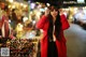 A woman in a red coat and sunglasses standing in front of a market.