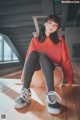 A woman sitting on a wooden floor wearing a red sweatshirt and black tights.